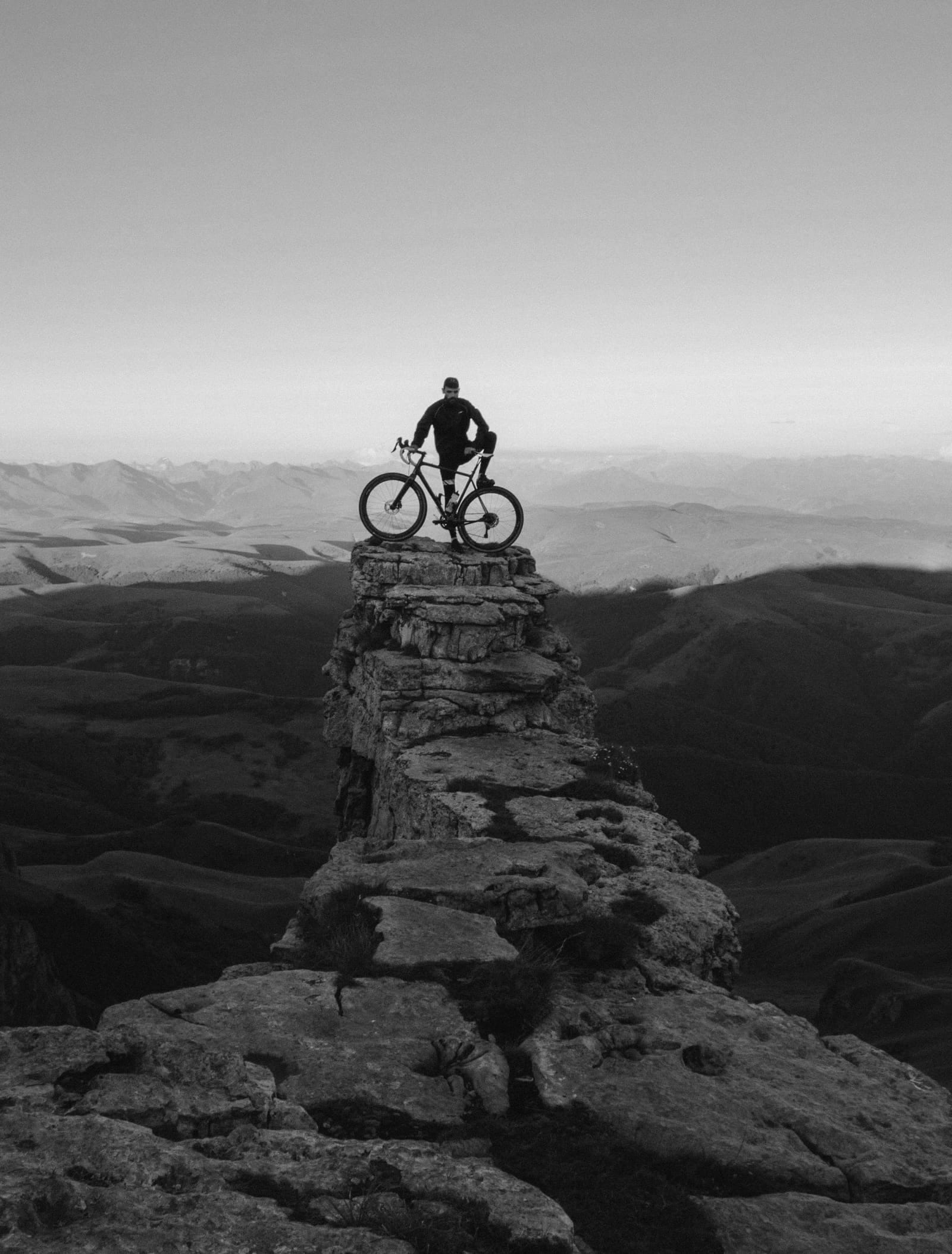 Cycliste immobile au sommet d'un promontoire rocheux, vélo à l'arrêt, face à un horizon de montagnes